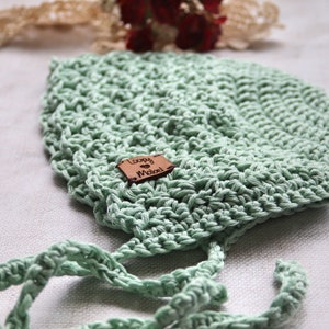 May include: A light green, crocheted baby bonnet with tie strings. The bonnet has a textured stitch pattern and a small, brown leather tag that reads "Loopy Mabel". The bonnet is made of soft yarn and is designed to fit a baby's head.