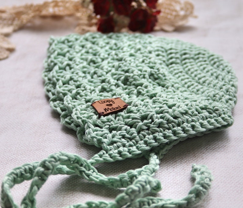 May include: A light green, crocheted baby bonnet with tie strings. The bonnet has a textured stitch pattern and a small, brown leather tag that reads "Loopy Mabel". The bonnet is made of soft yarn and is designed to fit a baby's head.