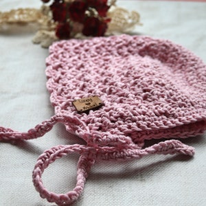 May include: A light pink, crocheted baby bonnet with a textured design. The bonnet has a small wooden tag that reads "Loopy Knots". The bonnet has attached ties for securing. The bonnet is displayed on a white surface.