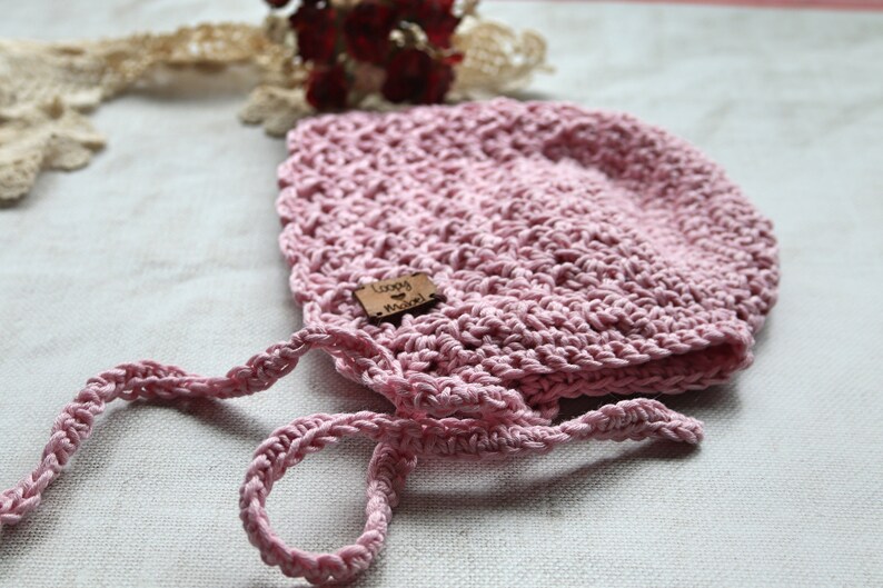 May include: A light pink, crocheted baby bonnet with a textured design. The bonnet has a small wooden tag that reads "Loopy Knots". The bonnet has attached ties for securing. The bonnet is displayed on a white surface.