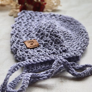 May include: A light purple, crocheted baby bonnet with tie strings. A small, square, brown leather tag is sewn on the bonnet with the words "Loopy Mabel" engraved on it. The bonnet is made of a soft, textured yarn.