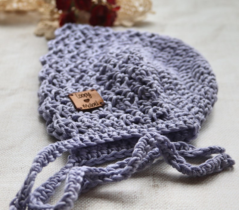 May include: A light purple, crocheted baby bonnet with tie strings. A small, square, brown leather tag is sewn on the bonnet with the words "Loopy Mabel" engraved on it. The bonnet is made of a soft, textured yarn.