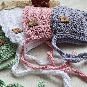 May include: Four handmade crochet bonnets in pastel colors: green, white, pink, and gray. Each bonnet has a matching tie and a small leather tag that reads "Loopy Mabel". The bonnets are arranged on a textured surface.