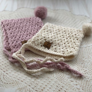 Op de afbeelding: Twee handgemaakte gehaakte baby mutsen. Eén is zacht crèmekleurig, de andere is licht paars. Elke muts heeft een bijpassende pompon en strikjes. De mutsen hebben een label met de tekst "Loopy & Mabel".