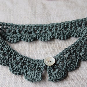 May include: A sage green, crocheted collar with a scalloped edge and a button closure. The collar is handmade and features a delicate, openwork design. The button is a pearlescent color.