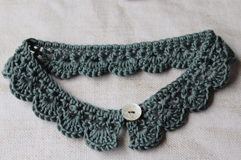 May include: A sage green, crocheted collar with a scalloped edge and a button closure. The collar is handmade and features a delicate, openwork design. The button is a pearlescent color.