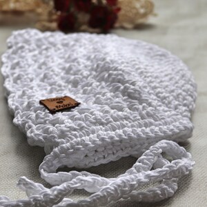 May include: A handmade white crochet bonnet with a scalloped edge and tie strings. A small brown leather tag is attached to the bonnet. The bonnet is designed for a baby or infant.