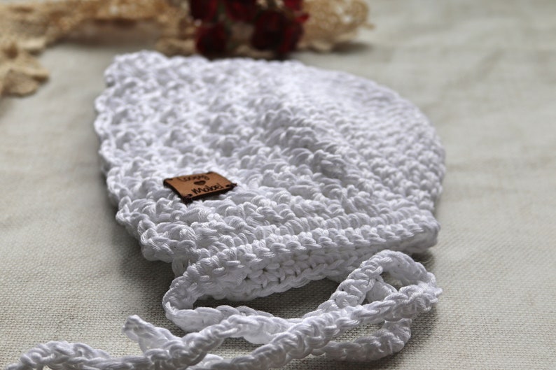 May include: A handmade white crochet bonnet with a scalloped edge and tie strings. A small brown leather tag is attached to the bonnet. The bonnet is designed for a baby or infant.