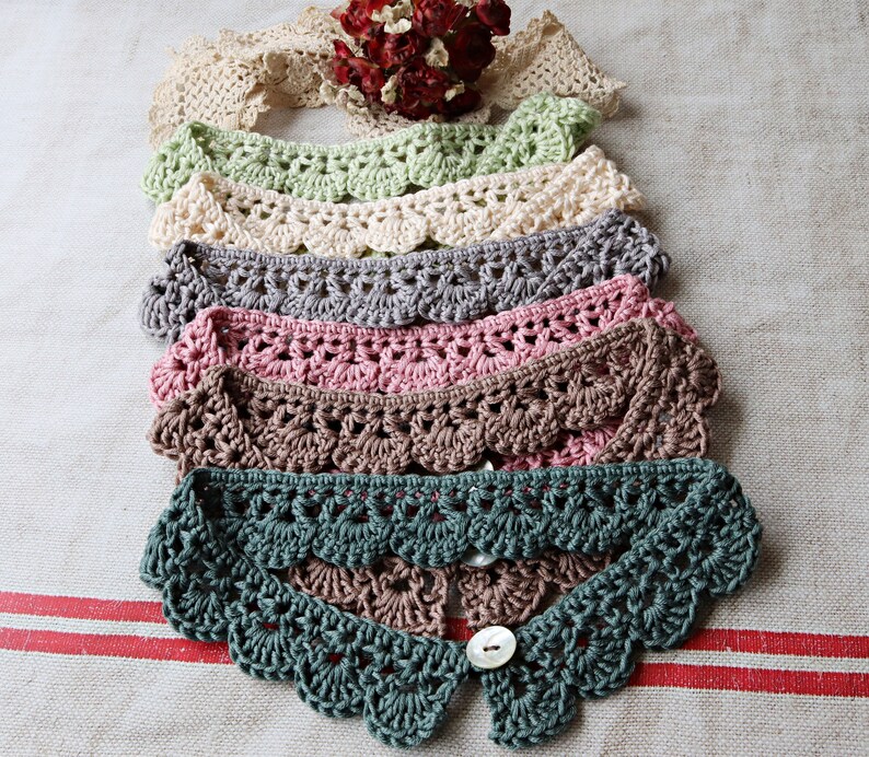 May include: A collection of handmade crochet collars in various colors, including green, cream, gray, pink, and brown. Each collar features a scalloped edge and a button closure. The collars are displayed on a neutral linen surface.