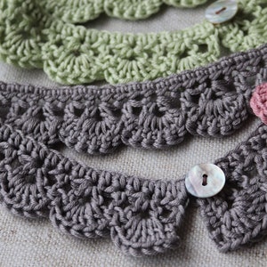 May include: Close-up of several crocheted lace collars in shades of gray, green, and pink. Each collar features a scalloped edge and is fastened with a button. The handmade collars are displayed on a neutral fabric background.