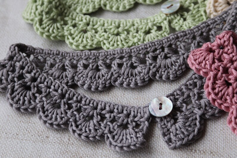 May include: Close-up of several crocheted lace collars in shades of gray, green, and pink. Each collar features a scalloped edge and is fastened with a button. The handmade collars are displayed on a neutral fabric background.