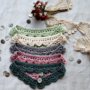 May include: A collection of five crocheted collars in various colors: light green, cream, gray, pink, and dark green. Each collar has a scalloped edge and a button closure. Decorative lace and buttons are also present.
