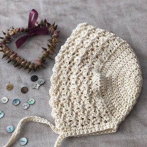 May include: Cream-colored, crocheted baby bonnet with a textured stitch pattern. The bonnet has a tie closure and is next to a heart-shaped dried flower wreath with a purple ribbon. Several buttons are scattered around.