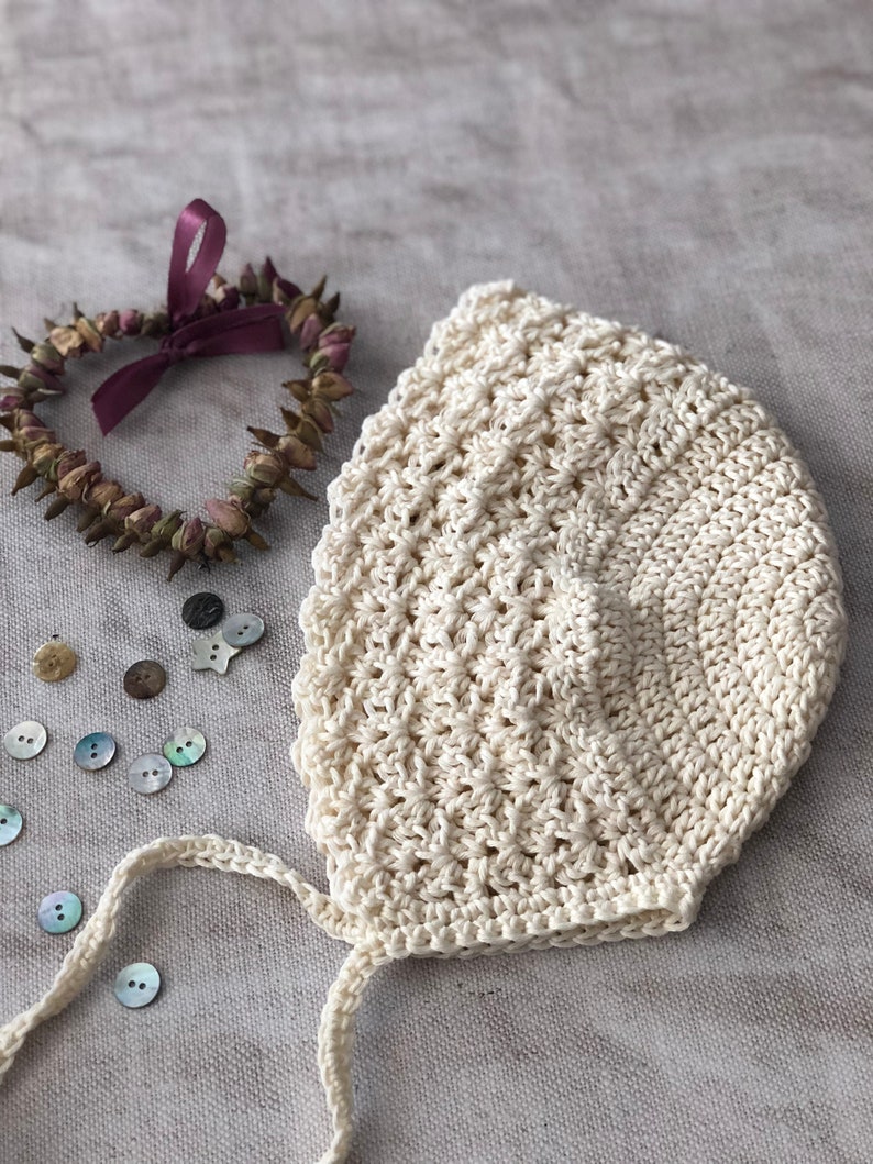 May include: Cream-colored, crocheted baby bonnet with a textured stitch pattern. The bonnet has a tie closure and is next to a heart-shaped dried flower wreath with a purple ribbon. Several buttons are scattered around.