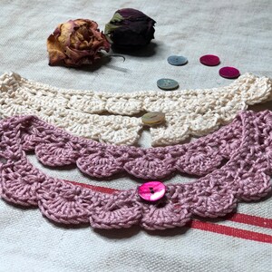 May include: Two crocheted collars in cream and mauve, each with a scalloped edge and a button closure. Dried rose buds and decorative buttons in various colors are scattered around the collars, suggesting a handmade craft item.