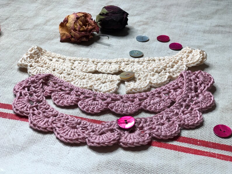 May include: Two crocheted collars in cream and mauve, each with a scalloped edge and a button closure. Dried rose buds and decorative buttons in various colors are scattered around the collars, suggesting a handmade craft item.