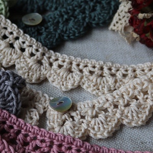 May include: Close-up of several crocheted lace trims in various colors, including cream, green, gray, and pink. Each trim features a scalloped edge and is adorned with a button. The handmade lace trims are suitable for craft projects.