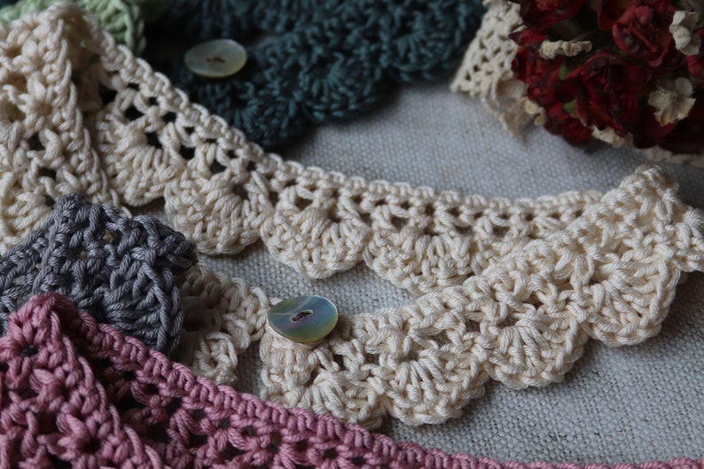 May include: Close-up of several crocheted lace trims in various colors, including cream, green, gray, and pink. Each trim features a scalloped edge and is adorned with a button. The handmade lace trims are suitable for craft projects.