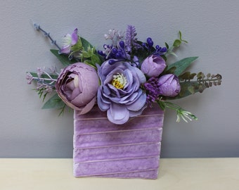 Op de afbeelding: Een paarse fluwelen vaas met een boeket paarse en lavendelkleurige bloemen. De bloemen zijn in een tros gerangschikt en omvatten rozen, lavendel en andere kleine paarse bloemen.