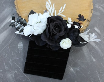 May include: A black velvet box adorned with a floral arrangement of black and white roses, white flowers, and dark green leaves. The arrangement sits on a light brown wooden slice, set against a gray background.