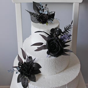 May include: Three-tiered white wedding cake decorated with black roses and greenery. The cake is adorned with black floral arrangements, including roses, leaves, and sprigs of lavender.