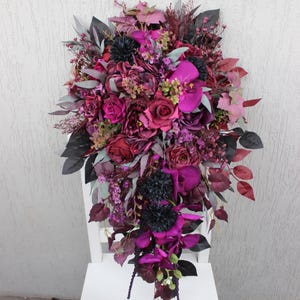 Könnte beinhalten: Ein kaskadenförmiges Blumenarrangement in Lila-, Magenta- und Burgundertönen. Der Strauß zeigt Rosen, Orchideen und andere Blumen mit dunkelgrünem und grauem Laub. Die Anordnung wird auf einem weißen Stuhl präsentiert.
