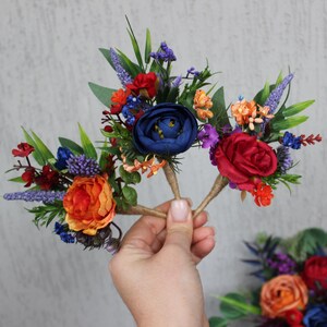 May include: Close-up of floral hair accessories featuring a vibrant mix of colors. The arrangement includes red, orange, blue, and purple flowers with green foliage. The accessories are held by a hand against a textured gray background.