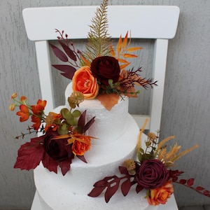 Può includere: Una torta bianca a tre piani decorata con fiori e foglie artificiali color borgogna e arancio. La torta è su una sedia bianca.