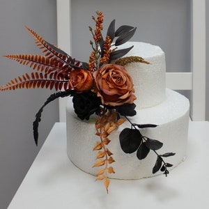 May include: Two white cake tiers decorated with two different floral arrangements. The first arrangement features a mix of orange, brown, and black flowers, while the second arrangement features a mix of orange, brown, and black flowers with a sunflower.