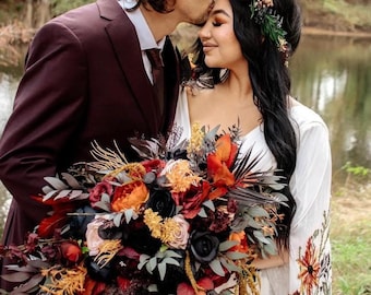 May include: A couple poses for a wedding photo. The groom is wearing a burgundy suit and kissing the bride on the forehead. The bride is wearing a white dress with floral embroidery and a floral crown. They are holding a large bouquet of dark red, orange, and black flowers.
