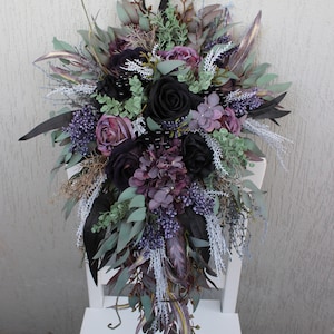 May include: A cascading floral arrangement featuring black roses, purple hydrangeas, and various greenery. The bouquet includes accents of lavender, sage, and dark leaves, creating a dramatic, elegant display. The arrangement is set against a white background.