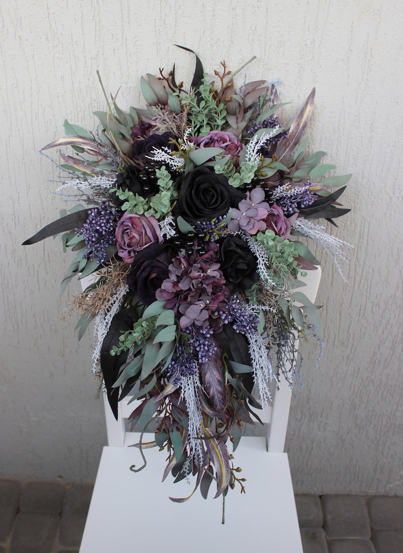 May include: A cascading floral arrangement featuring black roses, purple hydrangeas, and various greenery. The bouquet includes accents of lavender, sage, and dark leaves, creating a dramatic, elegant display. The arrangement is set against a white background.