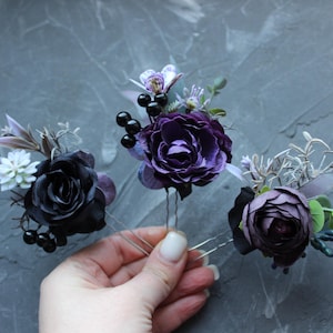 May include: Three floral hair pins featuring dark purple and black roses, berries, and greenery. The hair accessories are on silver-toned bobby pins. The flowers have a realistic appearance, with detailed petals and leaves. The background is a textured gray surface.