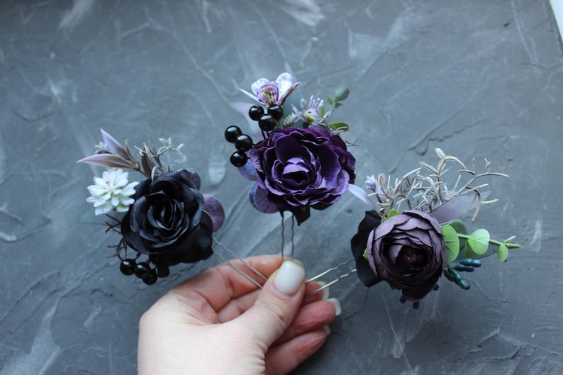 May include: Three floral hair pins featuring dark purple and black roses, berries, and greenery. The hair accessories are on silver-toned bobby pins. The flowers have a realistic appearance, with detailed petals and leaves. The background is a textured gray surface.