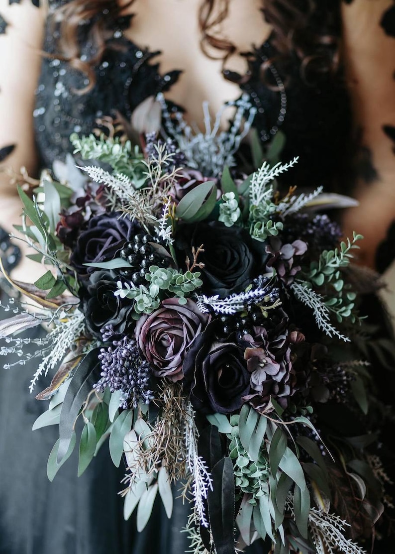 May include: A dark, moody bouquet featuring black and deep purple roses, accented with green foliage and delicate white sprigs. The arrangement includes berries and other textural elements, creating a gothic or alternative aesthetic. Perfect for a wedding or special event.