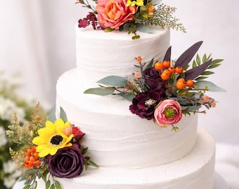 May include: A three-tiered white frosted cake decorated with colorful flowers. The flowers include yellow sunflowers, orange roses, and burgundy blooms. The cake sits on a wooden cake stand.