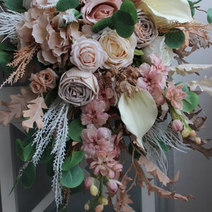 Puede incluir: Un ramo de flores artificiales en cascada de color rosa claro, crema y beige con vegetaci&oacute;n y hierbas secas. El ramo est&aacute; dispuesto en cascada y presenta rosas, hortensias y otros elementos florales.