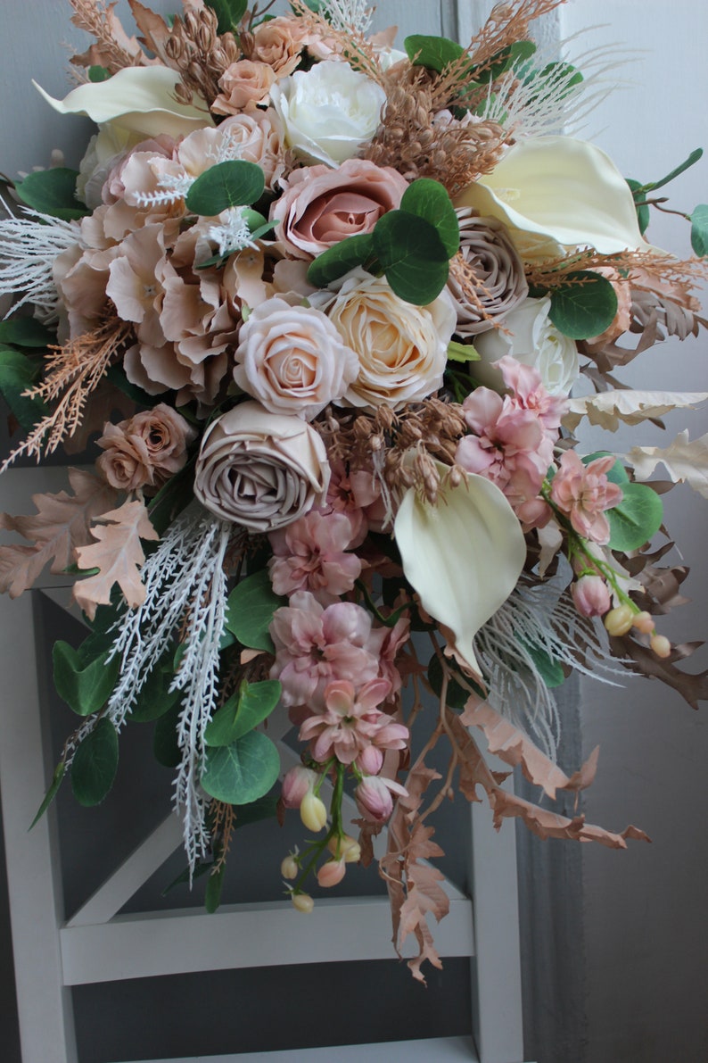 Puede incluir: Un ramo de flores artificiales en cascada de color rosa claro, crema y beige con vegetaci&oacute;n y hierbas secas. El ramo est&aacute; dispuesto en cascada y presenta rosas, hortensias y otros elementos florales.