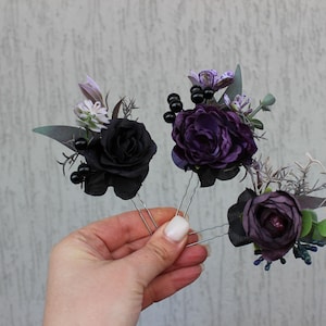 May include: Three floral hair pins featuring black and purple roses, accented with dark berries and greenery. The pins are held in a hand against a neutral background. These decorative hair accessories are ideal for special occasions.