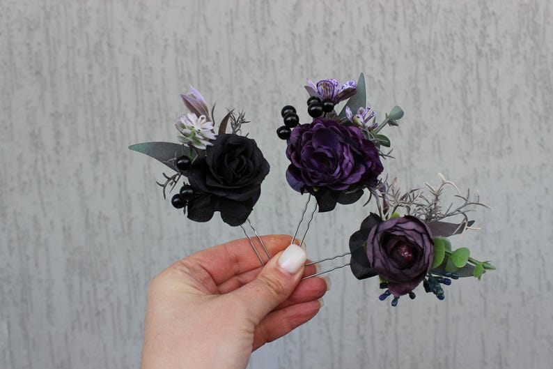 May include: Three floral hair pins featuring black and purple roses, accented with dark berries and greenery. The pins are held in a hand against a neutral background. These decorative hair accessories are ideal for special occasions.