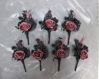May include: Eight burgundy rose boutonnieres with black berries and greenery. The boutonnieres have black stems and are arranged on a gray background.