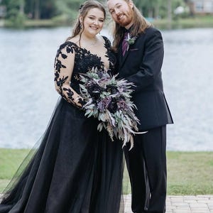 May include: A couple poses outdoors near a lake. The person on the left wears a black wedding gown with lace sleeves and a tulle skirt, holding a bouquet of purple flowers. The person on the right wears a black suit with a purple tie.