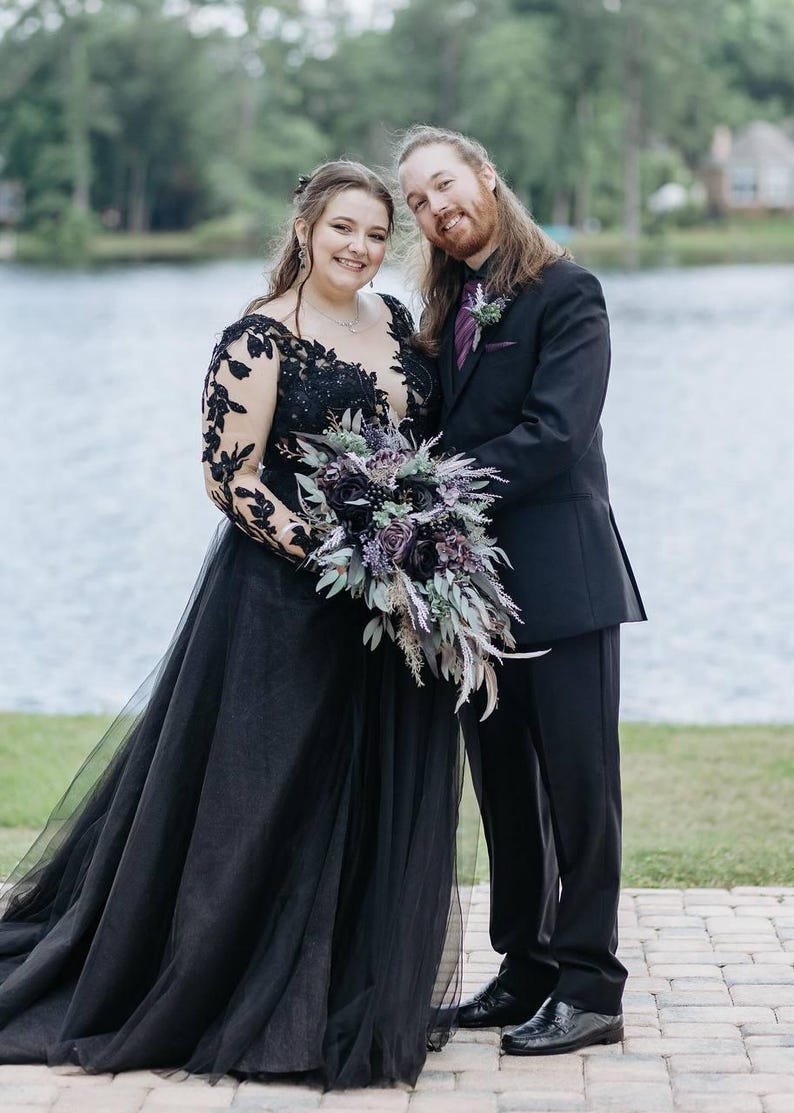 May include: A couple poses outdoors near a lake. The person on the left wears a black wedding gown with lace sleeves and a tulle skirt, holding a bouquet of purple flowers. The person on the right wears a black suit with a purple tie.