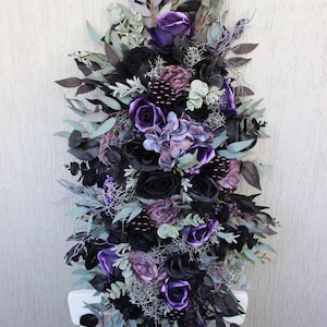 May include: A vertical floral arrangement featuring a mix of black, purple, and lavender artificial flowers and foliage. The arrangement includes roses, hydrangeas, and various leaves, creating a gothic or romantic aesthetic. The flowers are arranged on a white chair.