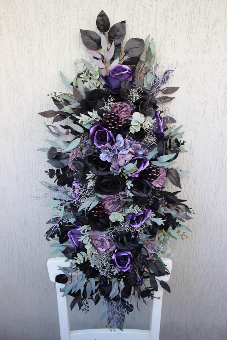 May include: A vertical floral arrangement featuring a mix of black, purple, and lavender artificial flowers and foliage. The arrangement includes roses, hydrangeas, and various leaves, creating a gothic or romantic aesthetic. The flowers are arranged on a white chair.
