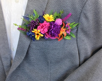 May include: A gray blazer with a colorful floral arrangement in the pocket. The arrangement includes yellow, pink, purple, and orange flowers with green foliage. A white dress shirt is visible underneath the blazer.
