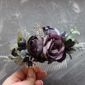 May include: A floral hair comb featuring dark purple roses and various greenery. The comb has a clear plastic base and is adorned with artificial flowers and foliage in shades of purple, green, and white.