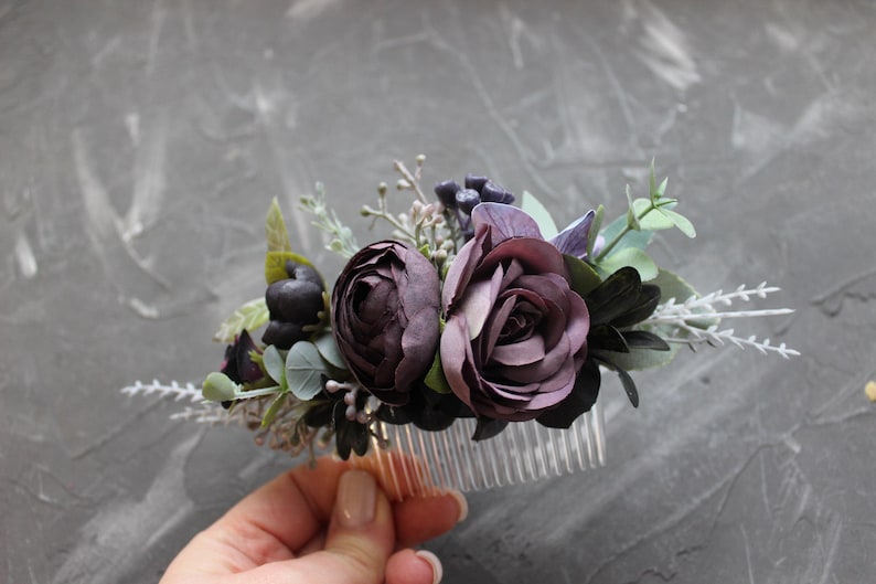 May include: A floral hair comb featuring dark purple roses and various greenery. The comb has a clear plastic base and is adorned with artificial flowers and foliage in shades of purple, green, and white.