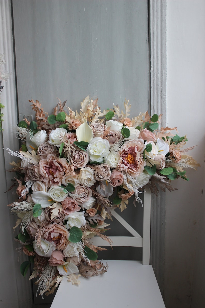 Puede incluir: Un gran arreglo floral con una mezcla de flores blancas, crema y rosa p&aacute;lido. El arreglo incluye rosas, peon&iacute;as y otras flores, as&iacute; como hierbas y hojas secas. El arreglo se muestra en una silla blanca.