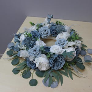 Könnte beinhalten: Ein kreisförmiger Blumenkranz mit künstlichen blauen, weißen und grünen Blumen und Blättern. Der Kranz enthält Rosen, Pfingstrosen und Eukalyptusblätter und schafft ein natürliches Design. Ideal für die Inneneinrichtung oder besondere Anlässe.
